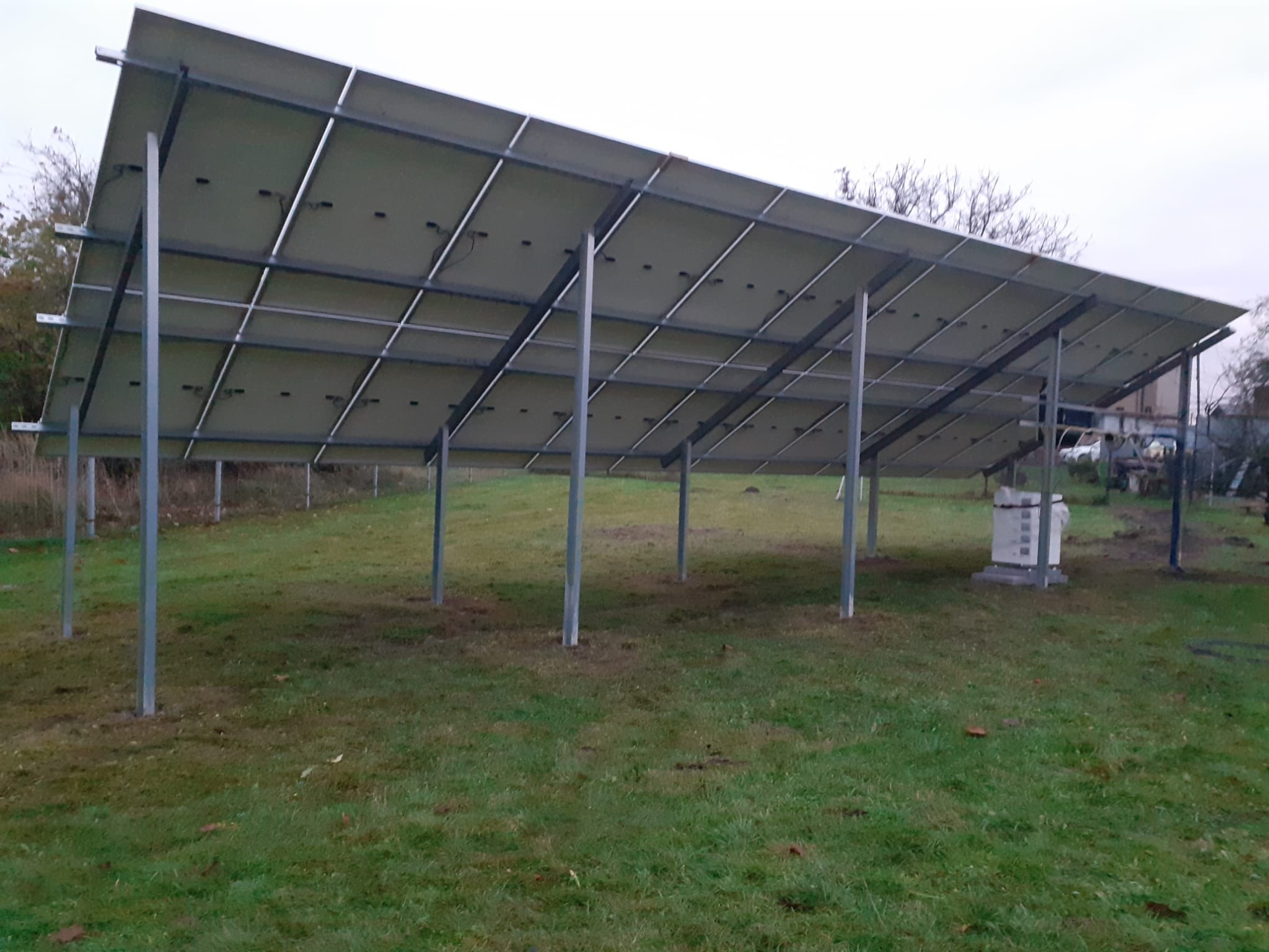 Widok od spodu na konstrukcję wsporczą paneli słonecznych, stalowe słupy na trawiastym terenie, w tle biała skrzynka elektryczna.