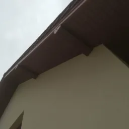 Fragment elewacji budynku z widocznym ociepleniem i brązowym podsufitką, sfotografowany pod kątem w pochmurny dzień.