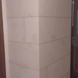 Wnętrze z narożnikiem wykończonym płytami imitującymi kamień, widoczne otwory na instalacje, narzędzia i fragment podłogi.