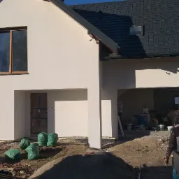 Nowo otynkowany dom jednorodzinny z ciemną dachówką, widoczne worki z materiałem budowlanym i pracownik w tle garażu.