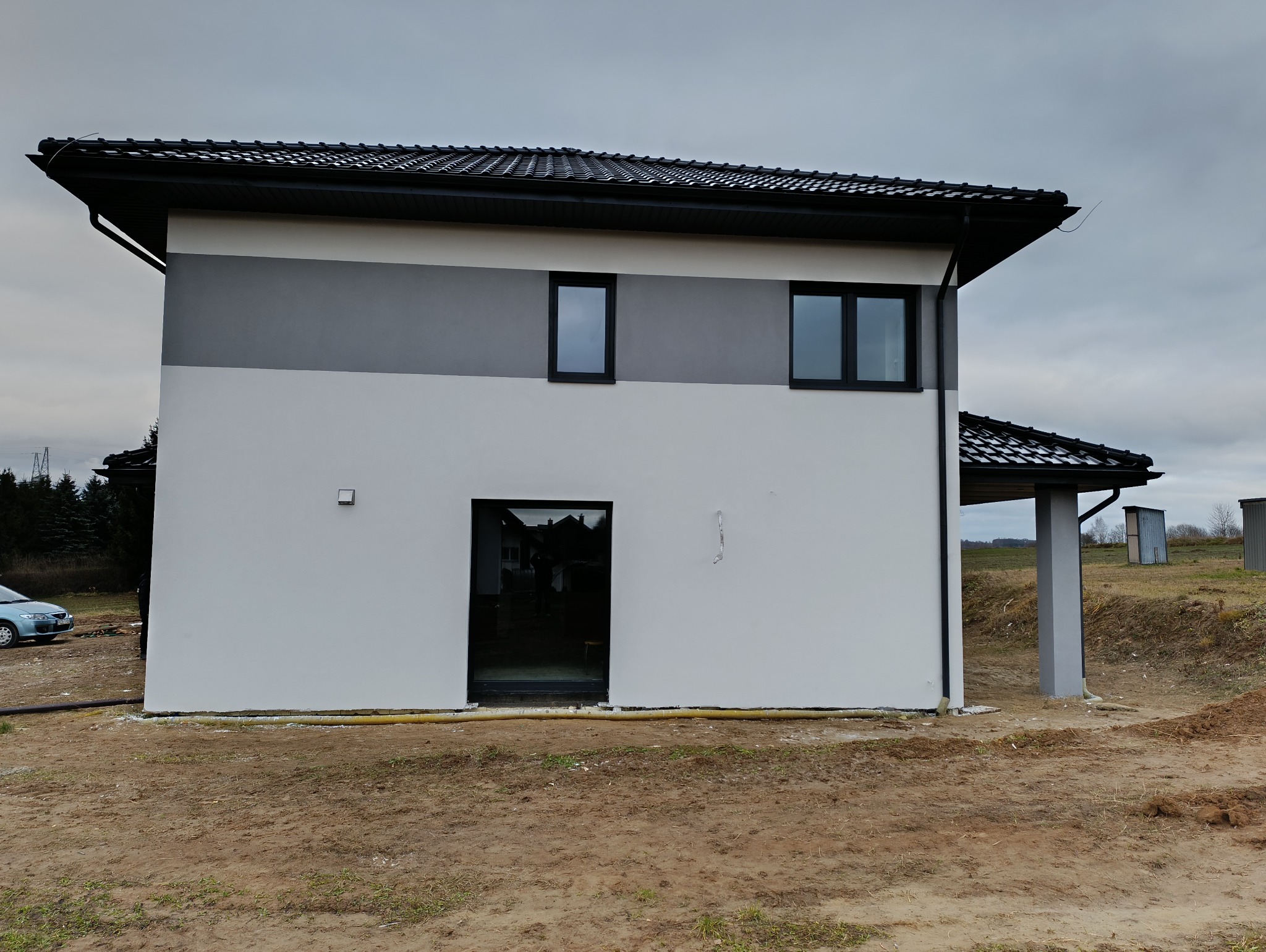 Nowoczesny dom z elewacją w odcieniach szarości i bieli, czarną stolarką okienną i dachem z ciemnej dachówki, widok z boku na tle pochmurnego nieba i przygotowanego terenu.