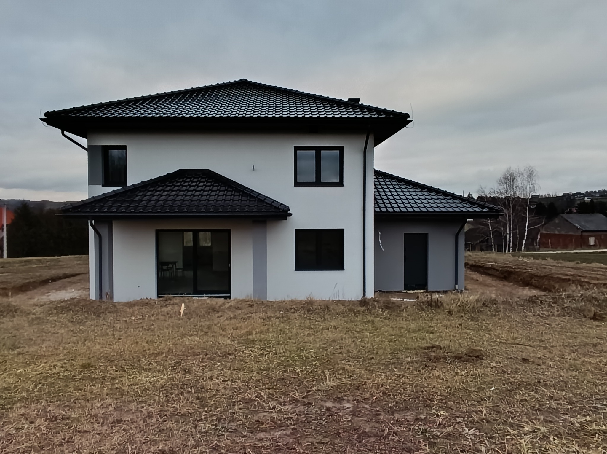 Dwukondygnacyjny dom z szarą elewacją i czarnym dachem z dachówki, widoczny z zewnątrz na tle pochmurnego nieba i jesiennego krajobrazu.