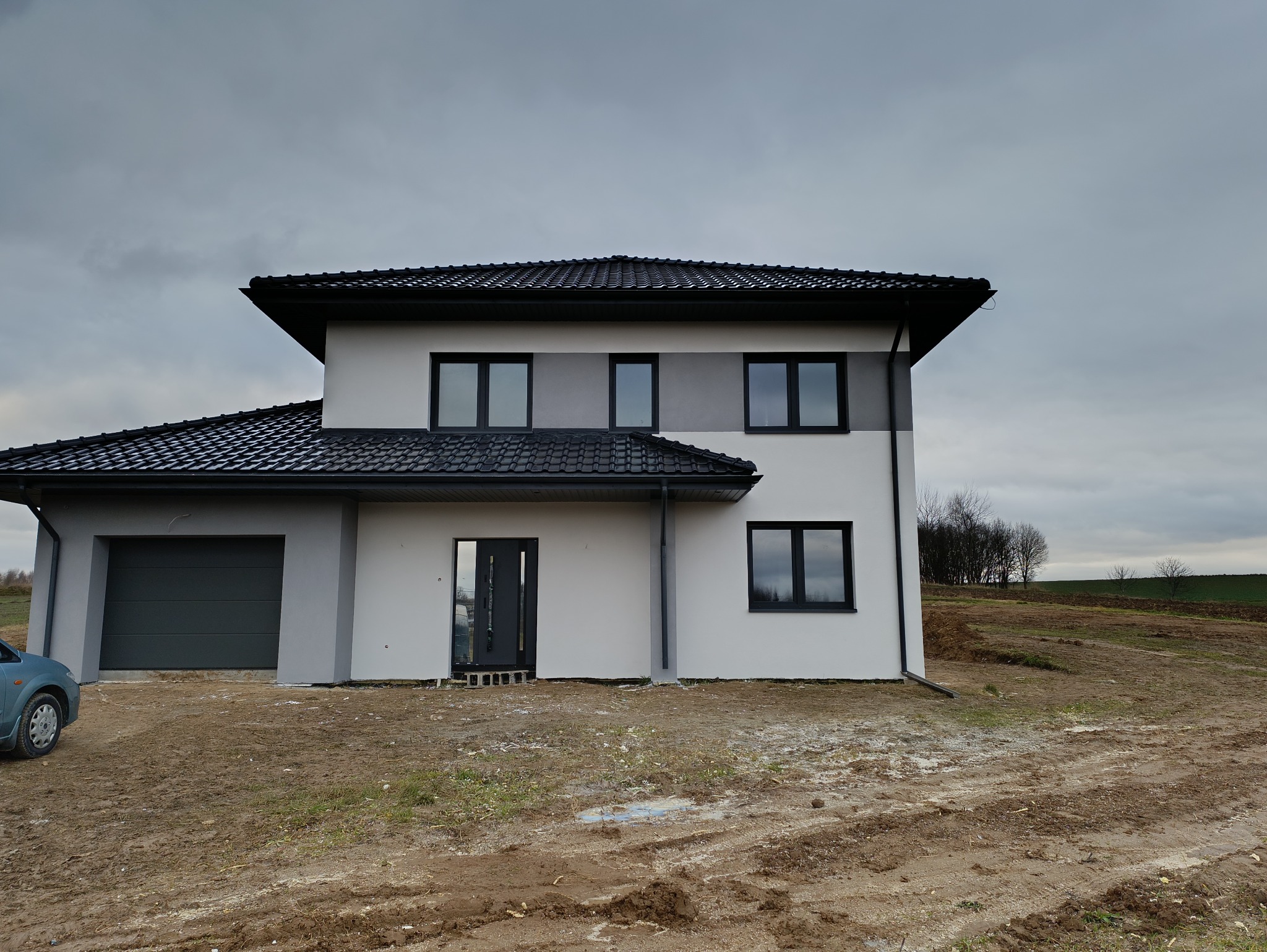 Nowoczesny dom jednorodzinny z szarą elewacją i czarną dachówką, widok z zewnątrz na tle pochmurnego nieba i zaoranego pola.