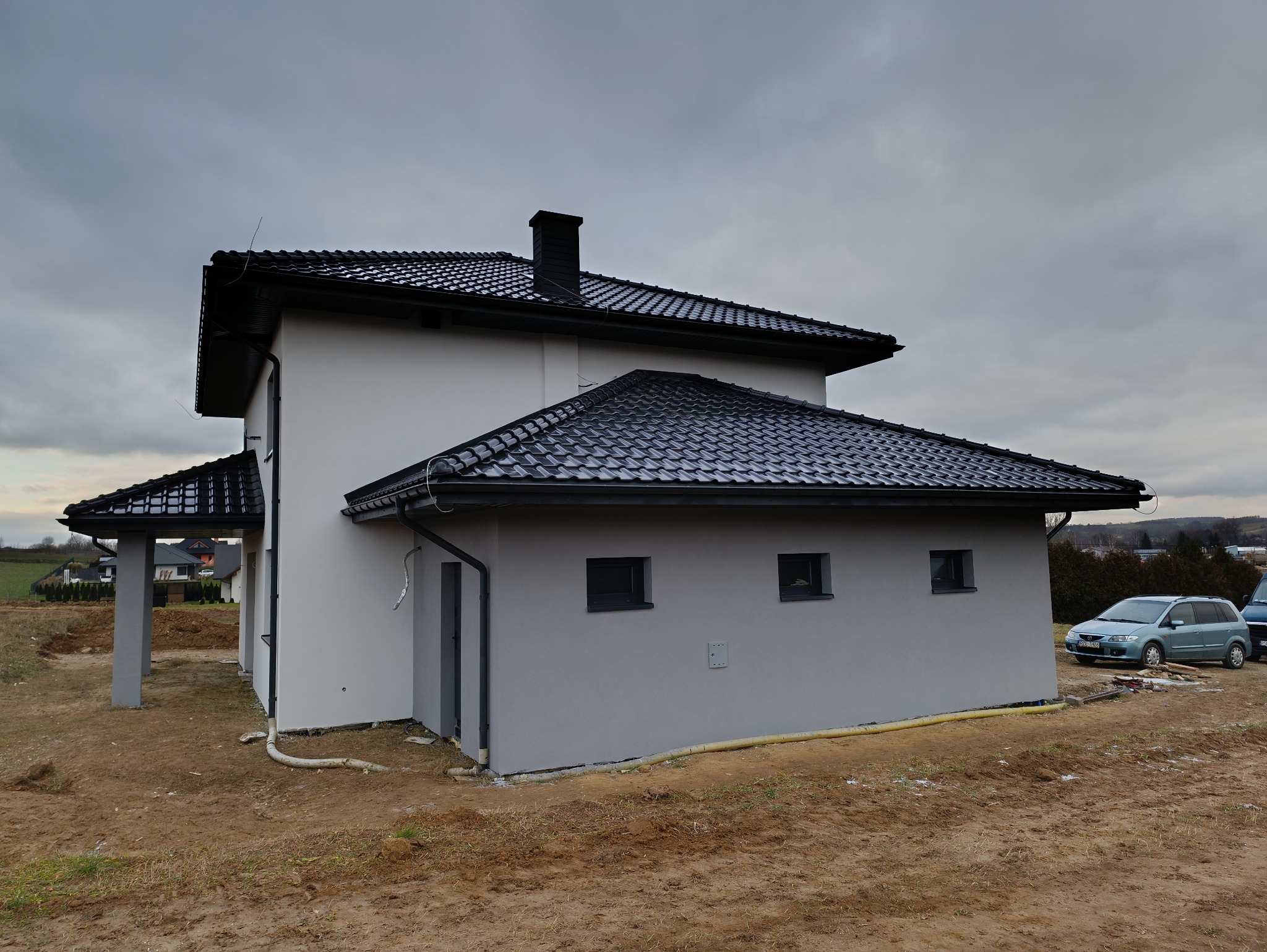 Nowo wybudowany dom z jasną elewacją i czarną dachówką, widok z boku na tle pochmurnego nieba, z widocznymi elementami wykończeniowymi i fragmentem terenu.