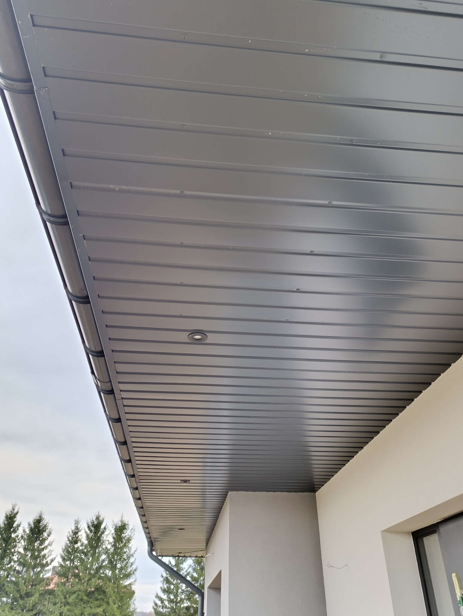 Szary podbitka dachowa z oświetleniem punktowym, widok z dołu na tle drzew i pochmurnego nieba.