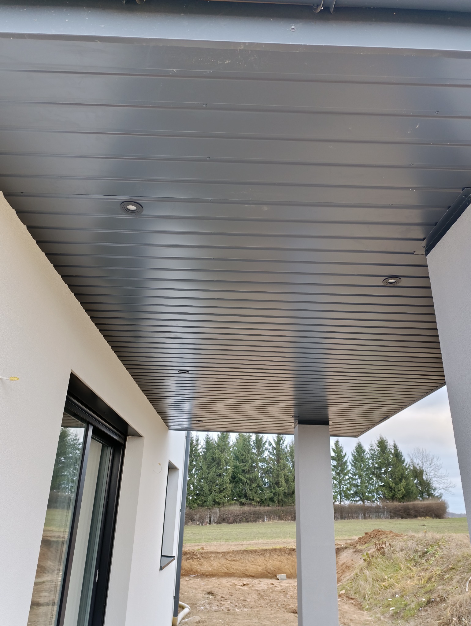 Podbitka dachowa z szarego panelu z wbudowanym oświetleniem punktowym, widok z dołu na tle zieleni drzew i wykopu ziemnego.