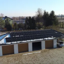 Nowoczesny garaż z trzema bramami i panelami słonecznymi na dachu, częściowo pokrytymi śniegiem, w zimowym krajobrazie.