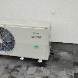 Zewnętrzna jednostka klimatyzacji Galmet Prima z widocznym symbolem R32, zamontowana na czarnych, gumowych podkładkach na szarej kostce brukowej przy białej ścianie budynku.