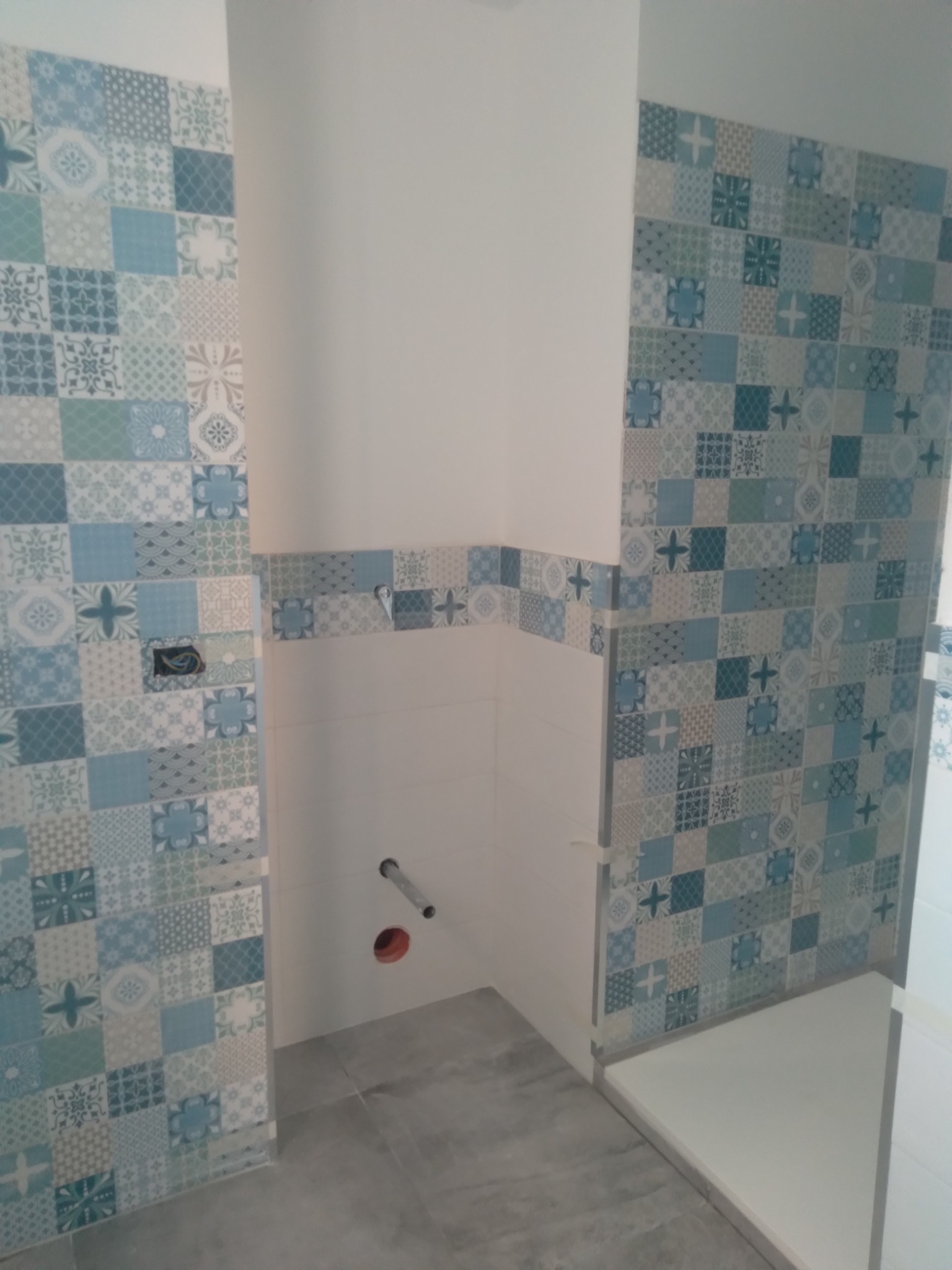 Niedokończona łazienka z patchworkowymi płytkami w odcieniach błękitu i szarości, widoczne wystające rury i otwór w ścianie, szara podłoga.