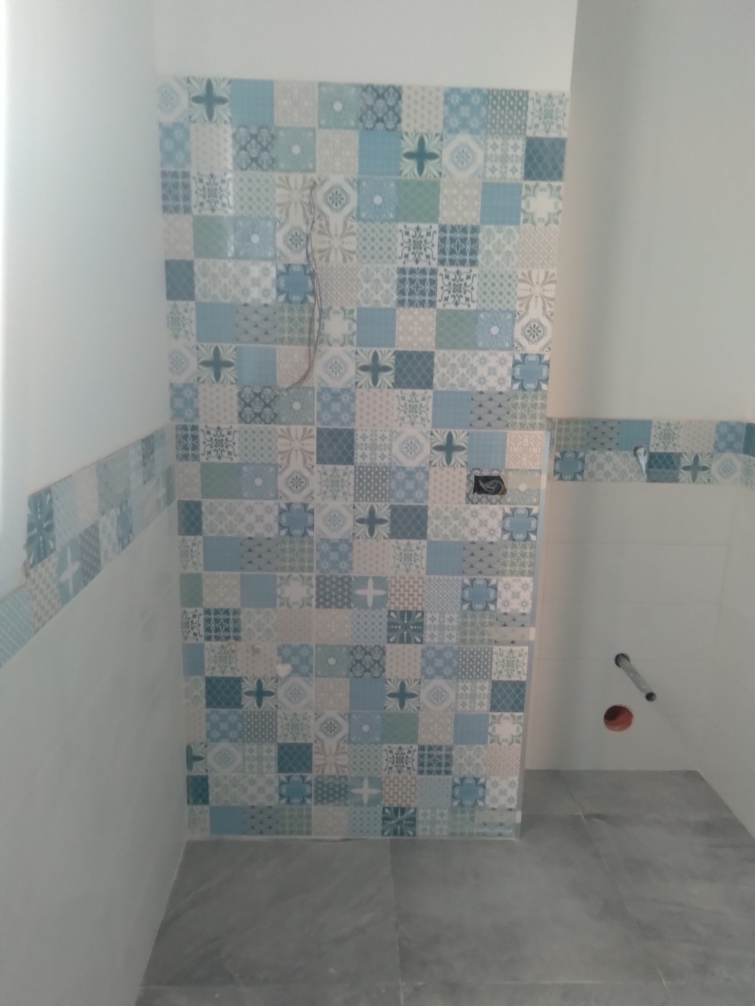 Ściana łazienki w trakcie wykańczania z niebiesko-białymi płytkami patchworkowymi, widoczne wystające kable i rury, szara podłoga.