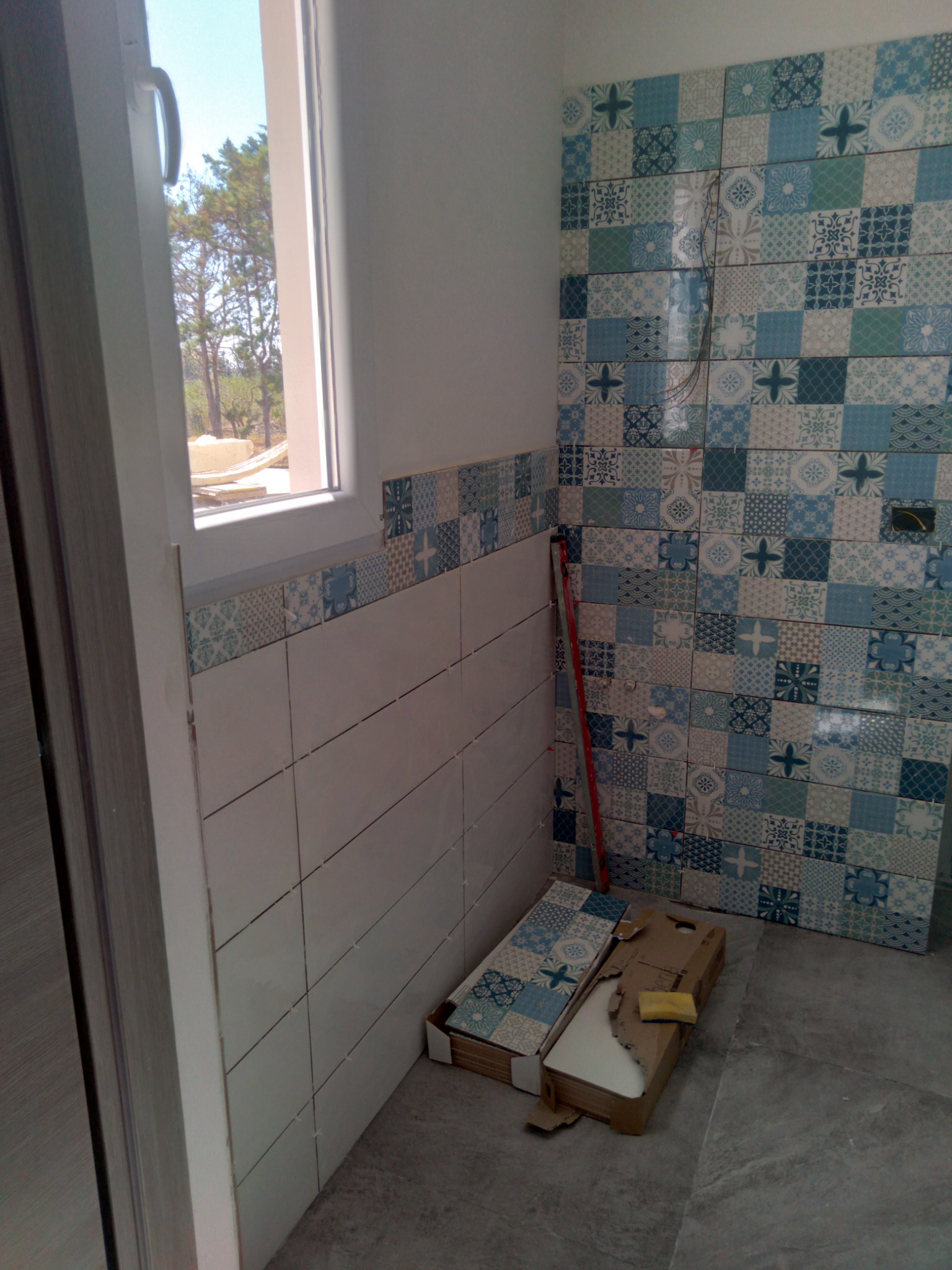 Wnętrze łazienki w trakcie układania płytek: dolna część ściany wyłożona białymi płytkami, górna część oraz fragment ściany z boku okna wyłożone dekoracyjnymi płytkami w odcieniach błękitu i bieli,...