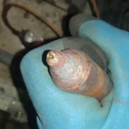 Miedziana rurka z lutem w niebieskiej rękawicy roboczej, widoczne ślady korozji i lutowania.