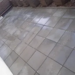 Szare, mokre płytki kostki brukowej po myciu, z widocznymi donicami z roślinami w tle.