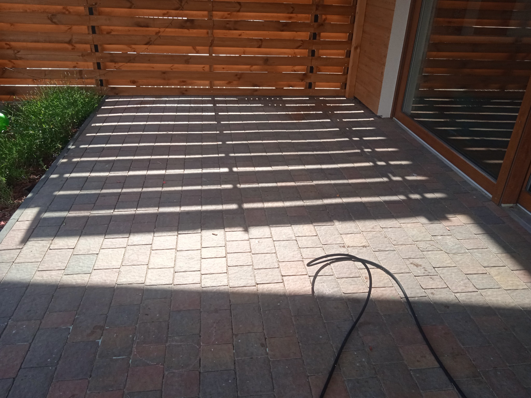 Ułożona kostka brukowa na tarasie z drewnianą ażurową ścianą w tle, rzucająca cienie w paski, z widocznym fragmentem węża ogrodowego w prawym dolnym rogu.
