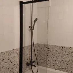 Nowoczesna kabina prysznicowa typu walk-in z czarnymi profilami i deszczownicą, zamontowana nad wanną z obudową z lastryko, białe płytki ścienne w tle.