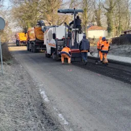 Frezowanie nawierzchni asfaltowej na drodze gminnej, ekipa w pomarańczowych kombinezonach, maszyna do frezowania i ciężarówki wywożące urobek.