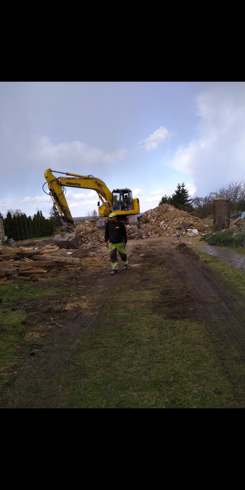 Żółta koparka Komatsu na placu budowy z gruzem, pracownik w odzieży roboczej przechadza się po terenie, widoczne pozostałości po rozebranym budynku.