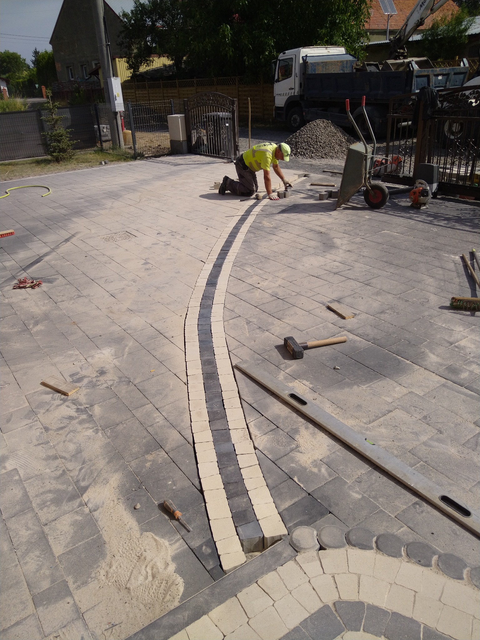 Pracownik w żółtej kamizelce układa mozaikę z kostki brukowej wzdłuż krętej ścieżki, widoczny samochód ciężarowy z wywrotką i sterta gruzu w tle.