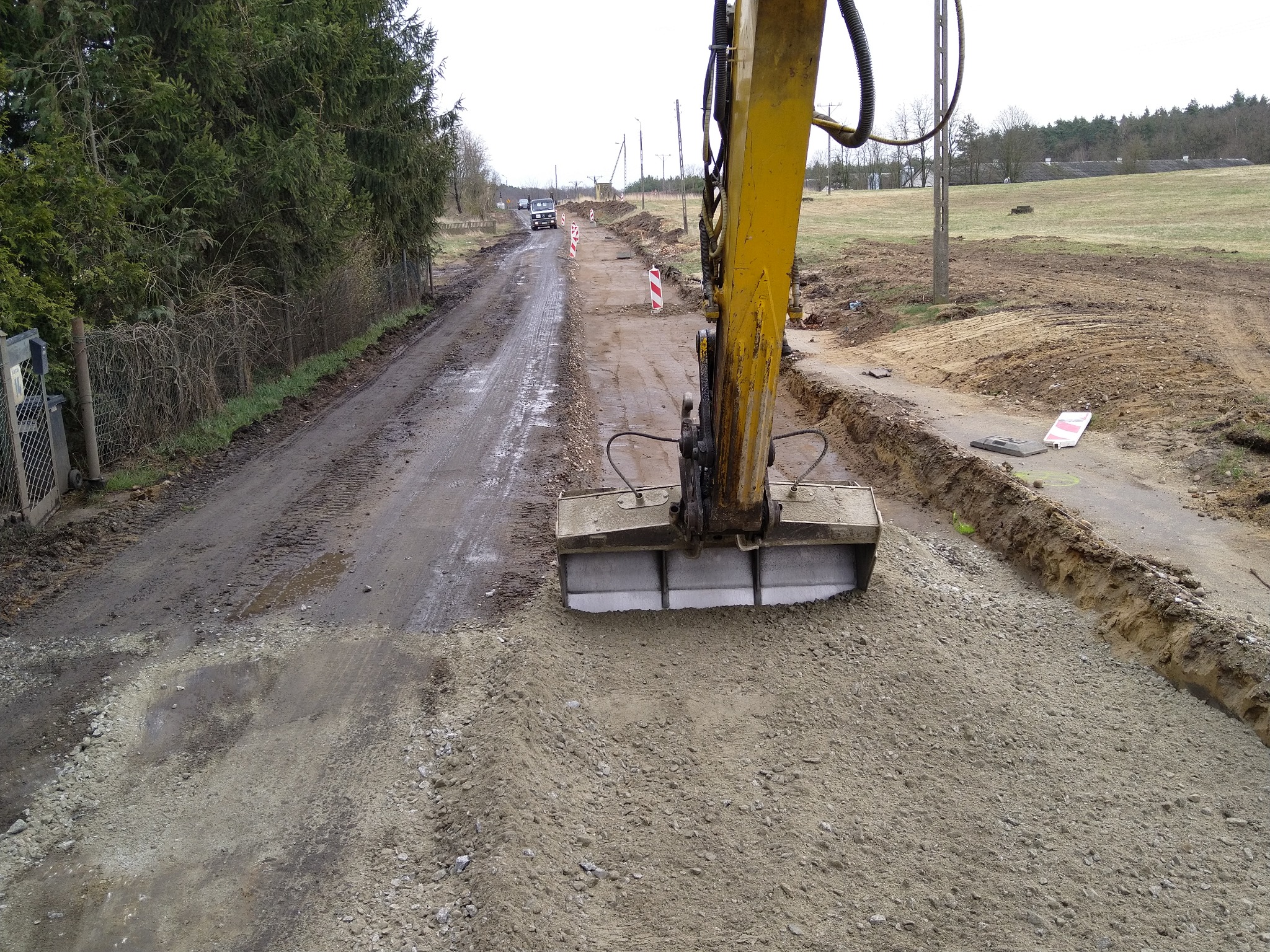 Żółta koparka równa teren pod budowę drogi, widoczne pachołki ostrzegawcze i wykopane rowy po bokach, w tle drzewa i pole.