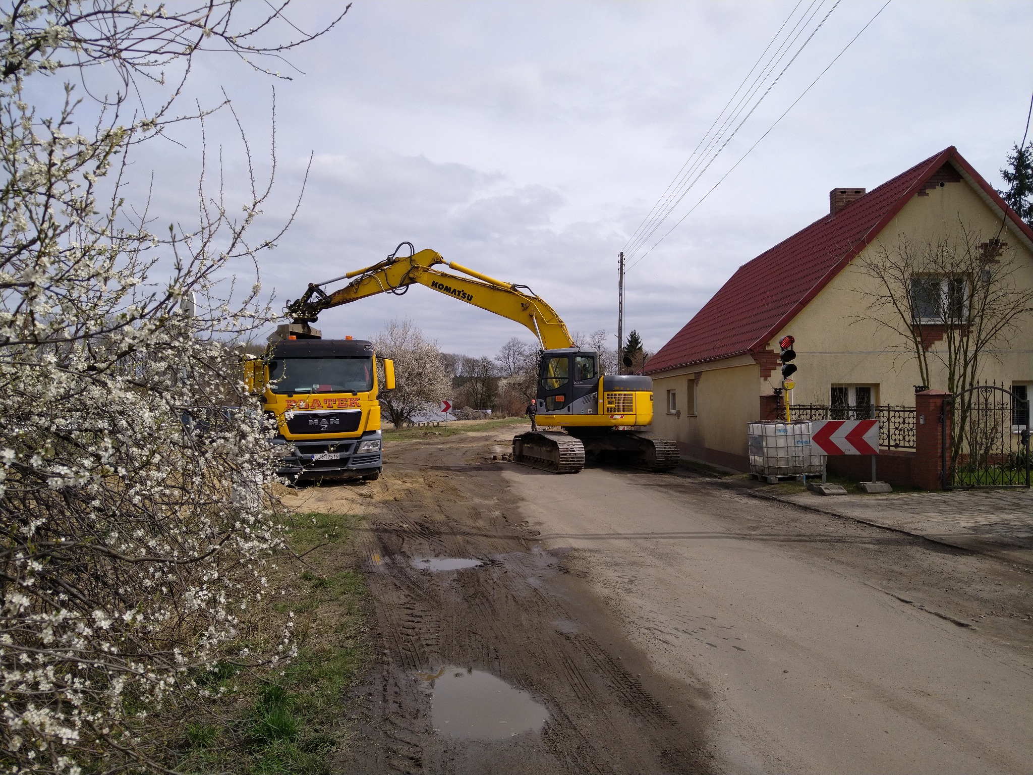 Żółta koparka Komatsu ładuje ziemię na żółtą ciężarówkę MAN na błotnistej drodze w pobliżu domu z czerwoną dachówką, z kwitnącym drzewem w pierwszym planie.