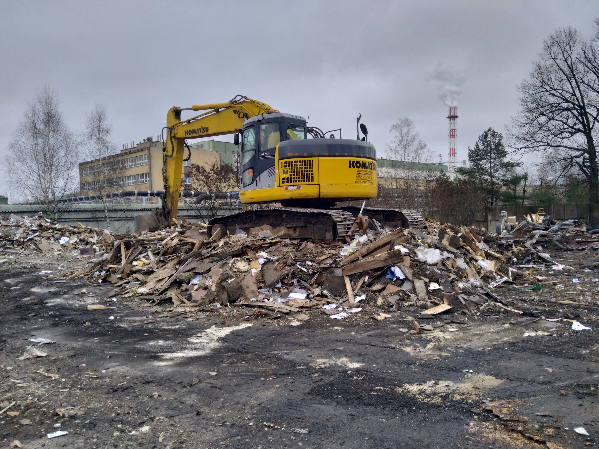 Żółta koparka Komatsu na stercie gruzu i odpadów po rozbiórce, w tle budynek i komin.