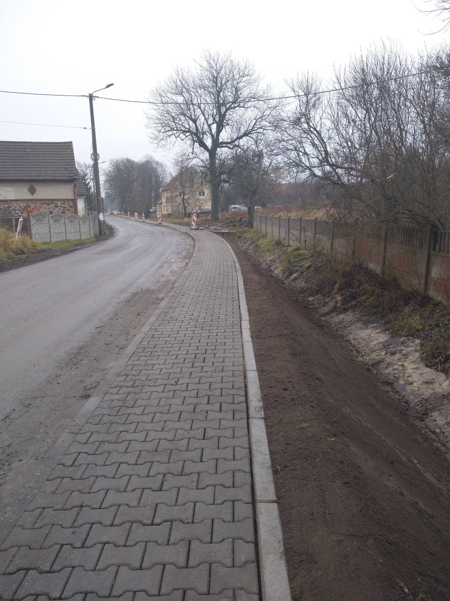 Ułożona szara kostka brukowa na chodniku wzdłuż drogi, z widocznym obrzeżem i świeżo wyrównanym pasem ziemi po prawej stronie, jesienny krajobraz w tle.