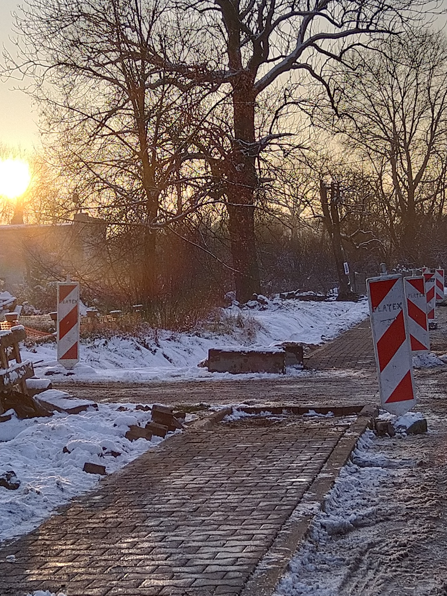 Świeżo ułożona kostka brukowa w zimowej scenerii, z widocznymi znakami ostrzegawczymi i fragmentami rozkopanej nawierzchni, oświetlona zachodzącym słońcem.