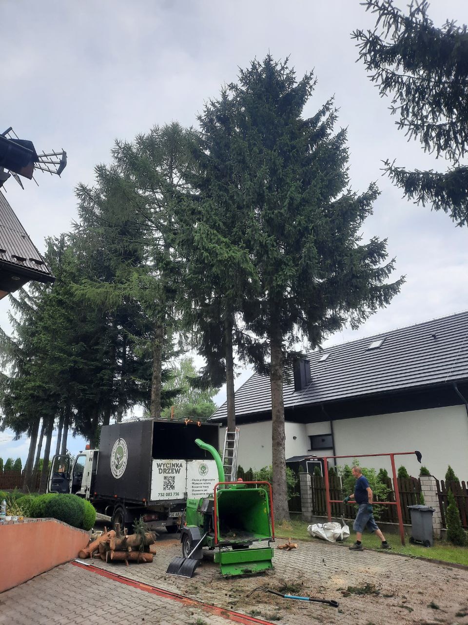 Ekipa z Krakowa przygotowuje się do usunięcia kilku wysokich świerków przed domem, z użyciem rębaka i samochodu z napisem 'Wycinka Drzew'.