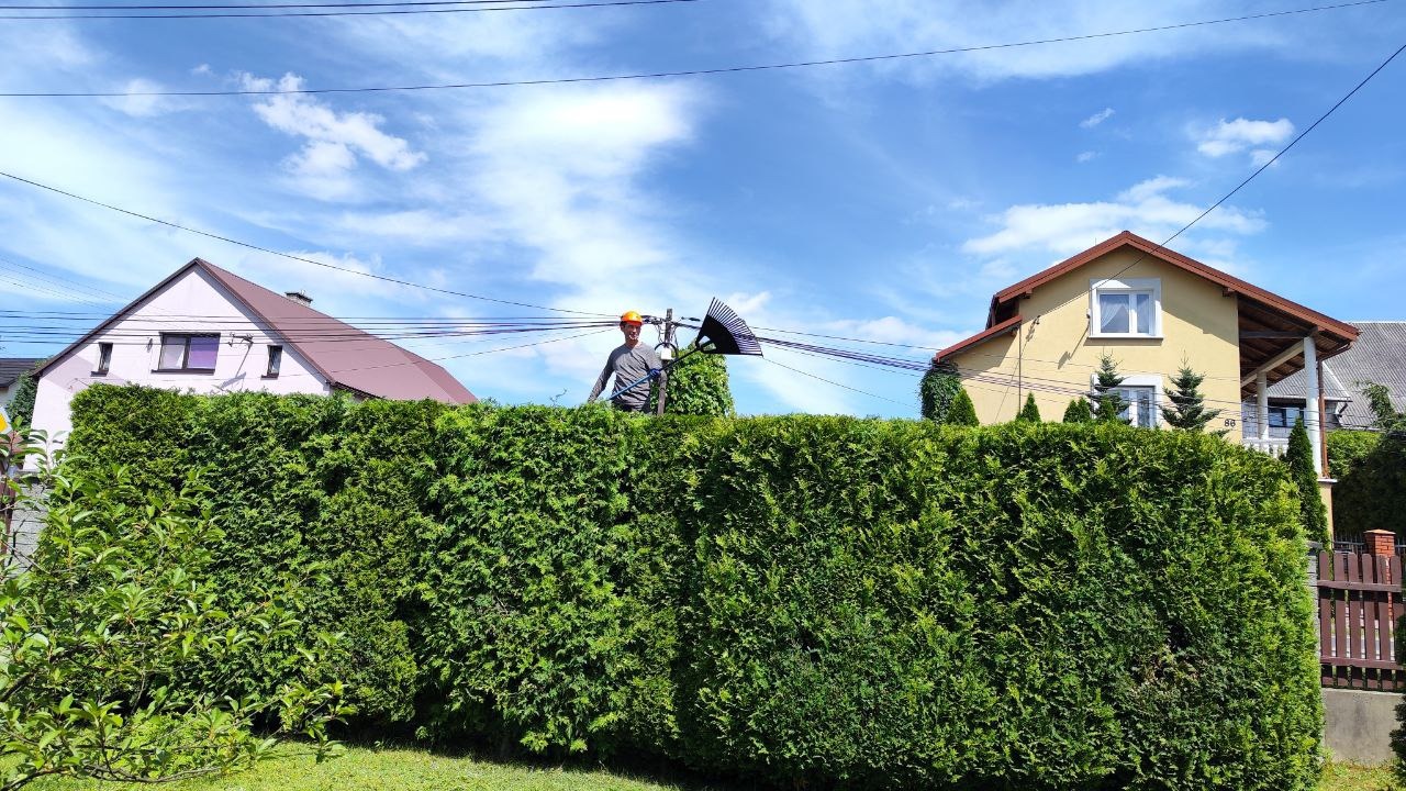 Mężczyzna w kasku, stojący za wysokim żywopłotem, trzyma długie nożyce do żywopłotu na tle domów i błękitnego nieba.