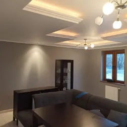 Szare wnętrze salonu z narożną kanapą, ciemnymi meblami i oświetleniem sufitowym, po świeżym odmalowaniu ścian.