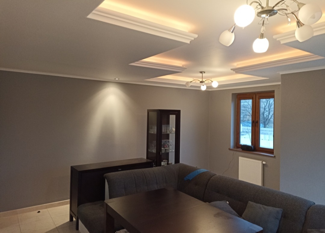 Szare wnętrze salonu z narożną kanapą, ciemnymi meblami i oświetleniem sufitowym, po świeżym odmalowaniu ścian.