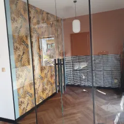 Nowoczesne, szklane drzwi przesuwne z czarną ramą, oddzielające pokój z wzorzystą tapetą w odcieniach złota i szarości od wnętrza z parkietem i ścianą w pepitkę.