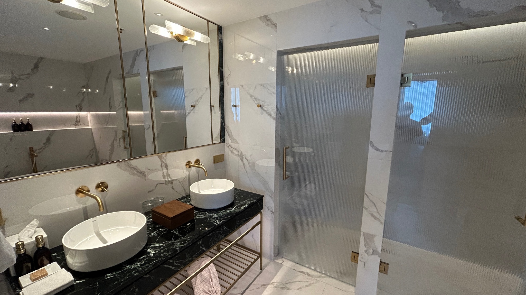 Elegancka łazienka z marmurowymi płytkami, umywalkami nablatowymi na czarnym blacie i kabiną prysznicową z ryflowanego szkła. Złote akcenty armatury i oświetlenia.