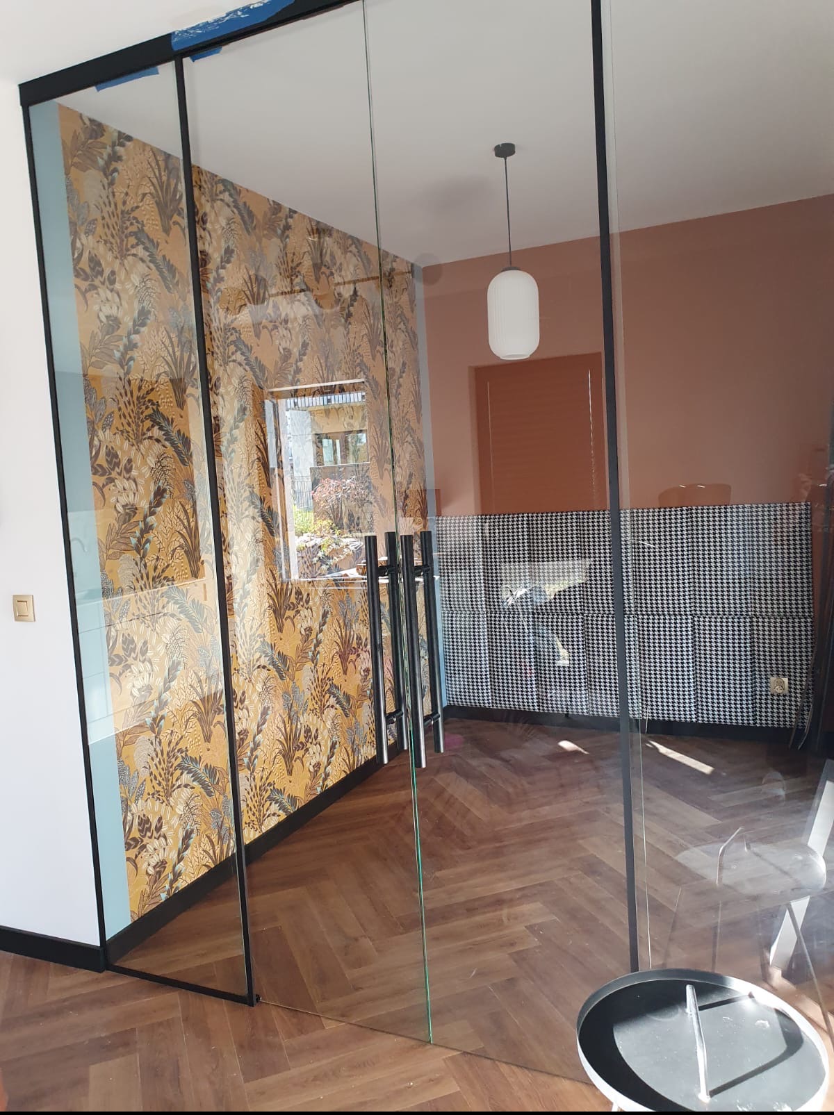 Nowoczesne, szklane drzwi przesuwne z czarną ramą, oddzielające pokój z wzorzystą tapetą w odcieniach złota i szarości od wnętrza z parkietem i ścianą w pepitkę.