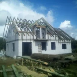 Dwukondygnacyjny budynek w stanie surowym otwartym, z widoczną konstrukcją dachu, wykonany z białych bloczków, z oknami bez szyb i otworami drzwiowymi, na tle błękitnego nieba z chmurami.