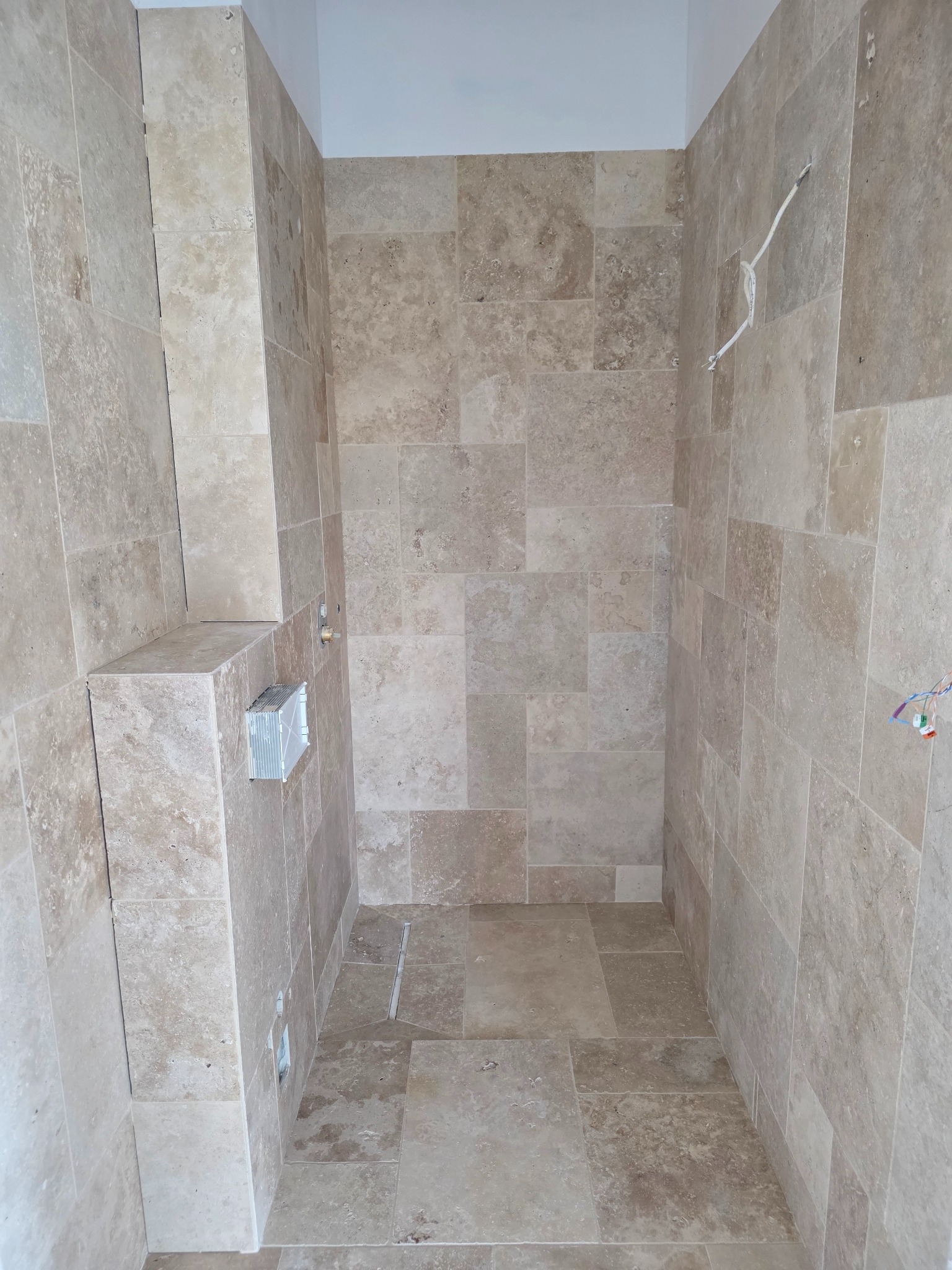 Wykończona łazienka z beżowymi płytkami trawertynowymi na ścianach i podłodze, widoczne elementy instalacji i otwór wentylacyjny.