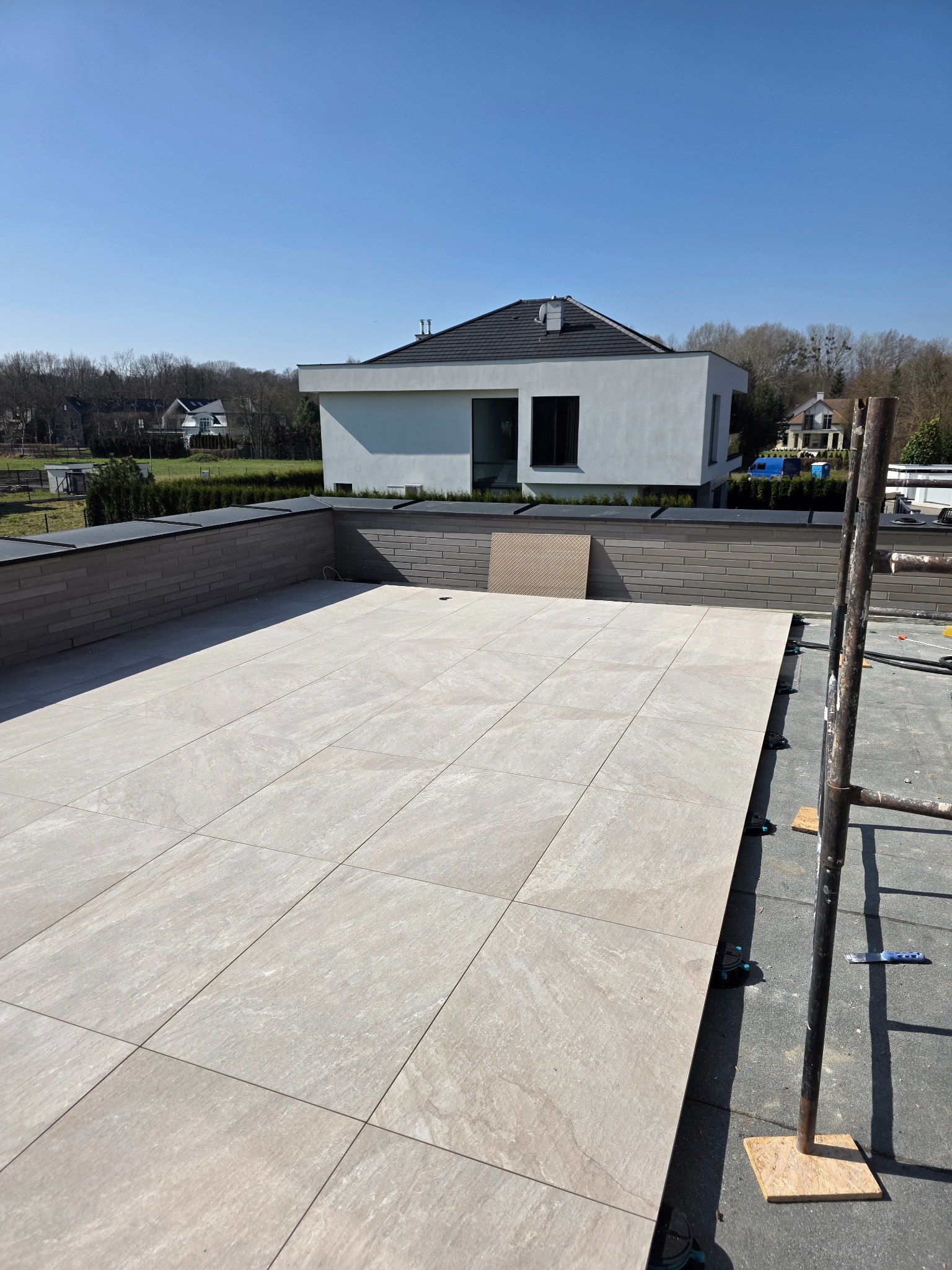 Jasne płytki tarasowe ułożone na dachu budynku, z widocznymi elementami konstrukcyjnymi i domem w tle, w słoneczny dzień. Układanie płytek na dachu.