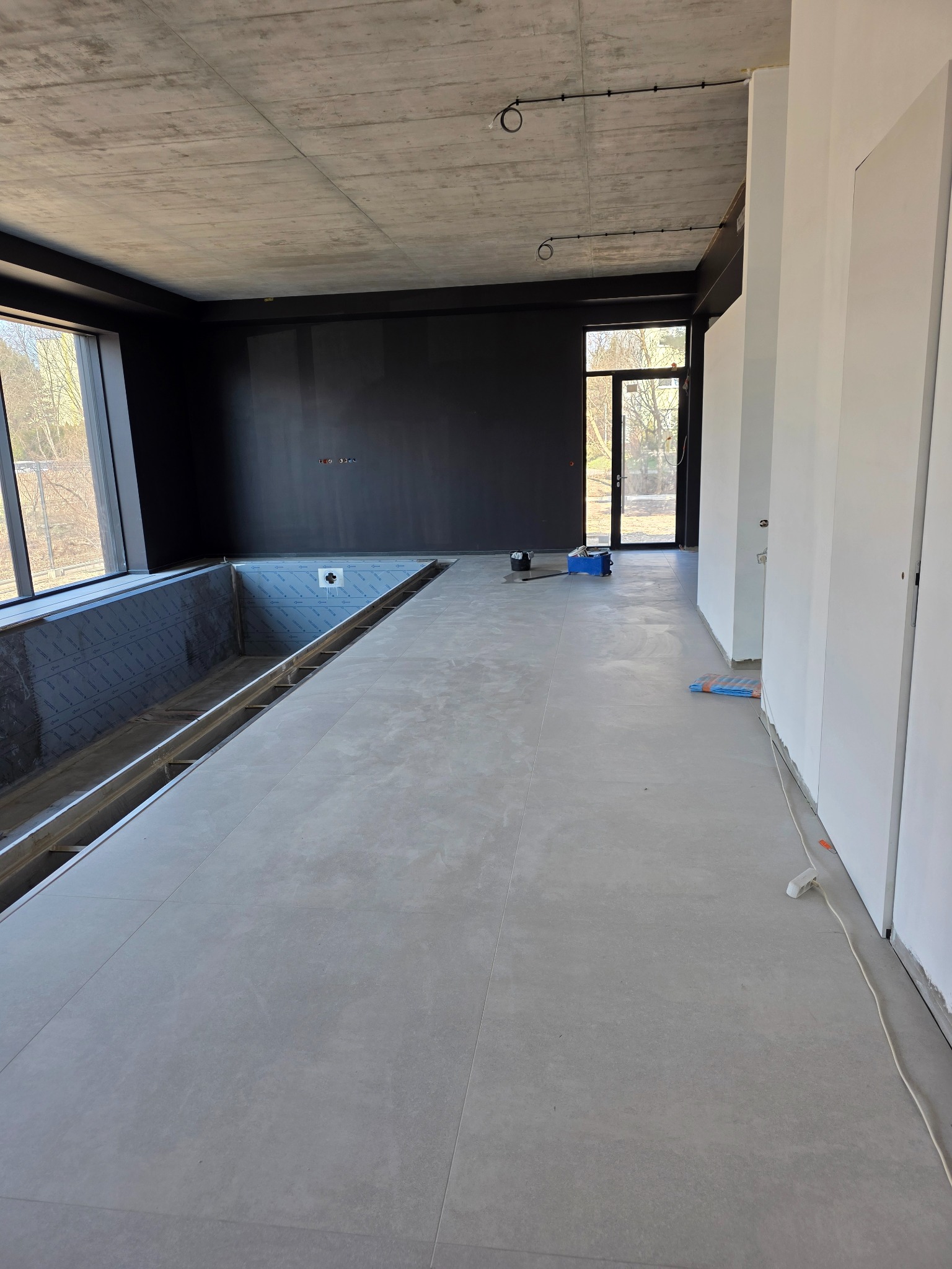 Wnętrze w trakcie wykończenia: szare płytki podłogowe, czarna ściana, surowy betonowy sufit z instalacją elektryczną. Widać przygotowany basen w pomieszczeniu.