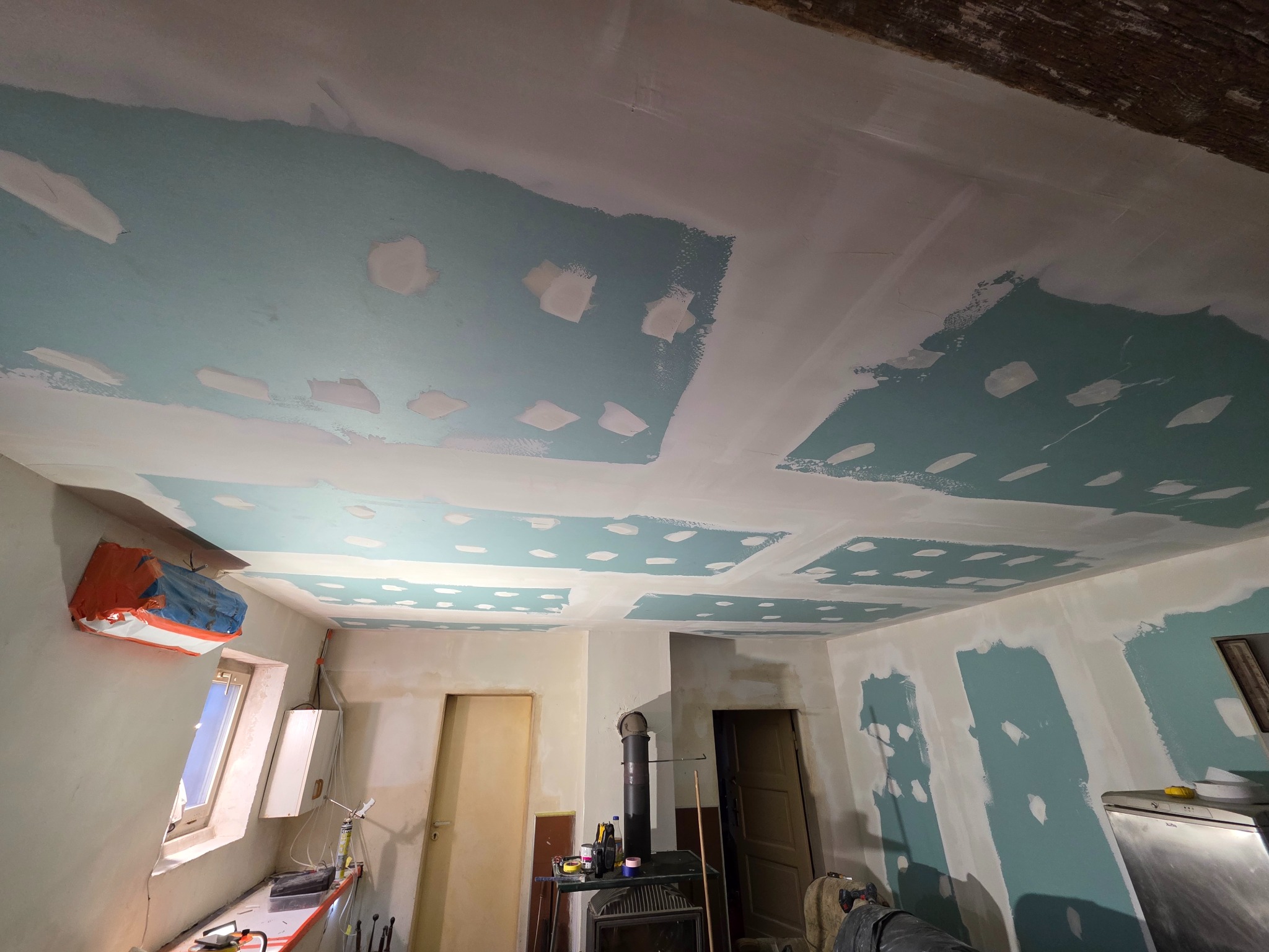 Pokój w trakcie remontu: ściany i sufit częściowo pomalowane na niebiesko, z widocznymi łatami szpachli. W tle piec i okno zabezpieczone folią malarską.