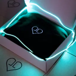 Otwarty karton z czarnym materiałem w środku, ozdobiony neonowym sznurkiem, na tle innych zamkniętych kartonów z logo firmy.