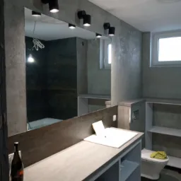 Nowoczesna łazienka w szarościach z lustrem podświetlonym czarnymi lampami punktowymi, z widoczną butelką wina na blacie umywalki i toaletą w tle.