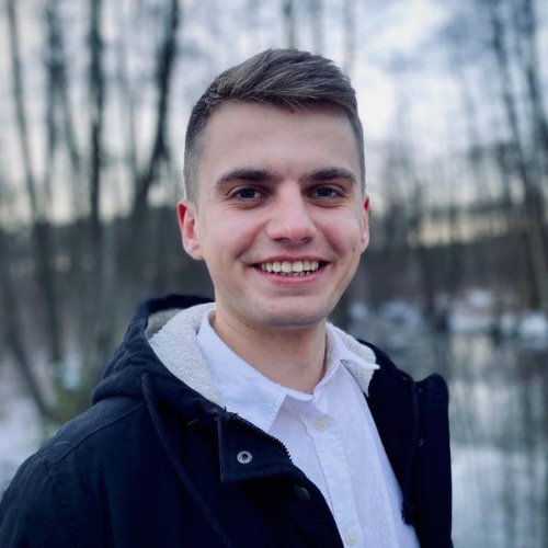 Portret uśmiechniętego mężczyzny w czarnej kurtce z białym kołnierzem, na tle zimowego krajobrazu z drzewami i śniegiem.