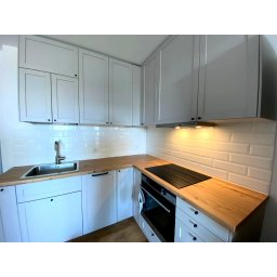 PRECYZJA Firma Remontowo-Budowlana - Jasna kuchnia z białymi szafkami, drewnianym blatem i płytkami imitującymi cegłę na ścianie.