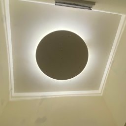 PRECYZJA Firma Remontowo-Budowlana - Nisko kątowe ujęcie sufitu z zamontowanym okrągłym panelem LED z delikatną poświatą, dekoracyjną listwą przysufitową i fragmentem ściany z widocznymi śladami po niedawnym malowaniu.