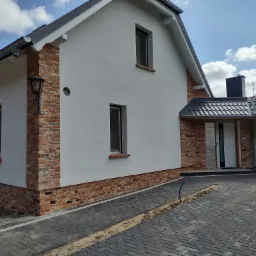 Nowoczesny dom jednorodzinny z białą elewacją i elementami z czerwonej cegły, widoczny fragment podjazdu z kostki brukowej w odcieniach szarości, błękitne niebo z chmurami w tle.