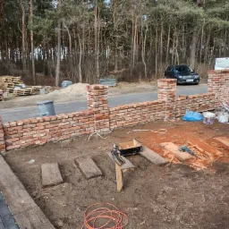 Budowa niskiego muru z czerwonej cegły w ogrodzie, widoczne narzędzia murarskie, palety z cegłami w tle, samochód osobowy zaparkowany przy drodze.