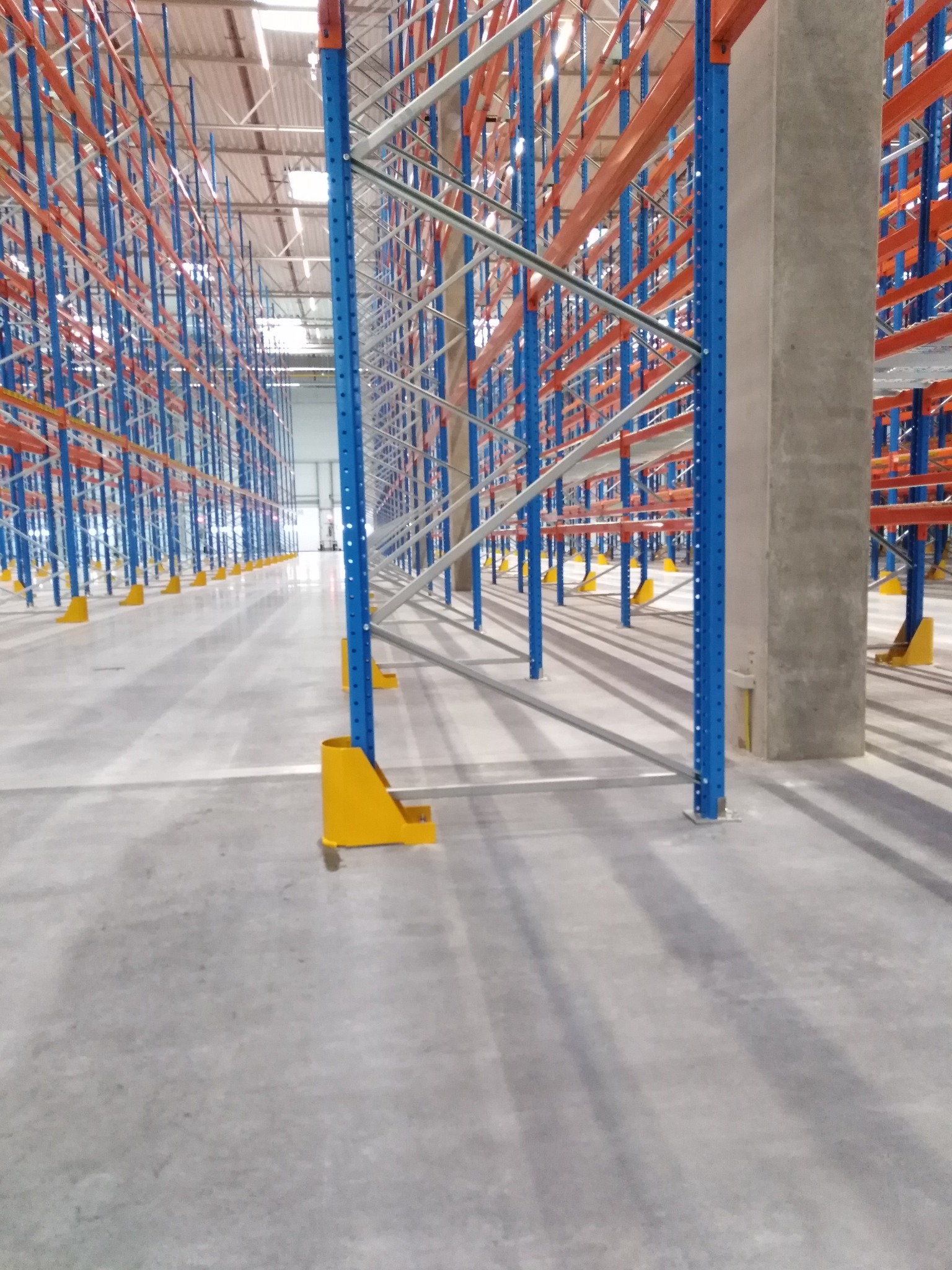 Pusta hala magazynowa z systemem regałów wysokiego składowania w kolorach niebieskim i pomarańczowym, betonowa podłoga z widocznymi śladami po czyszczeniu.