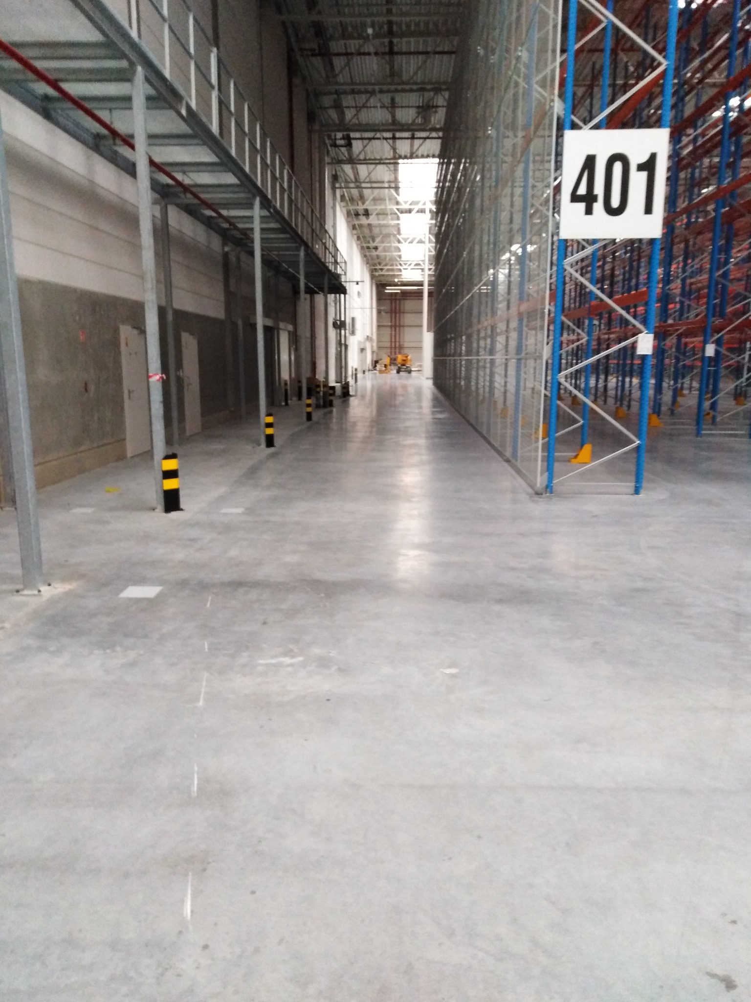 Pusta hala magazynowa z betonową podłogą, metalowymi regałami i numerem 401 na słupie, widok w głąb hali.