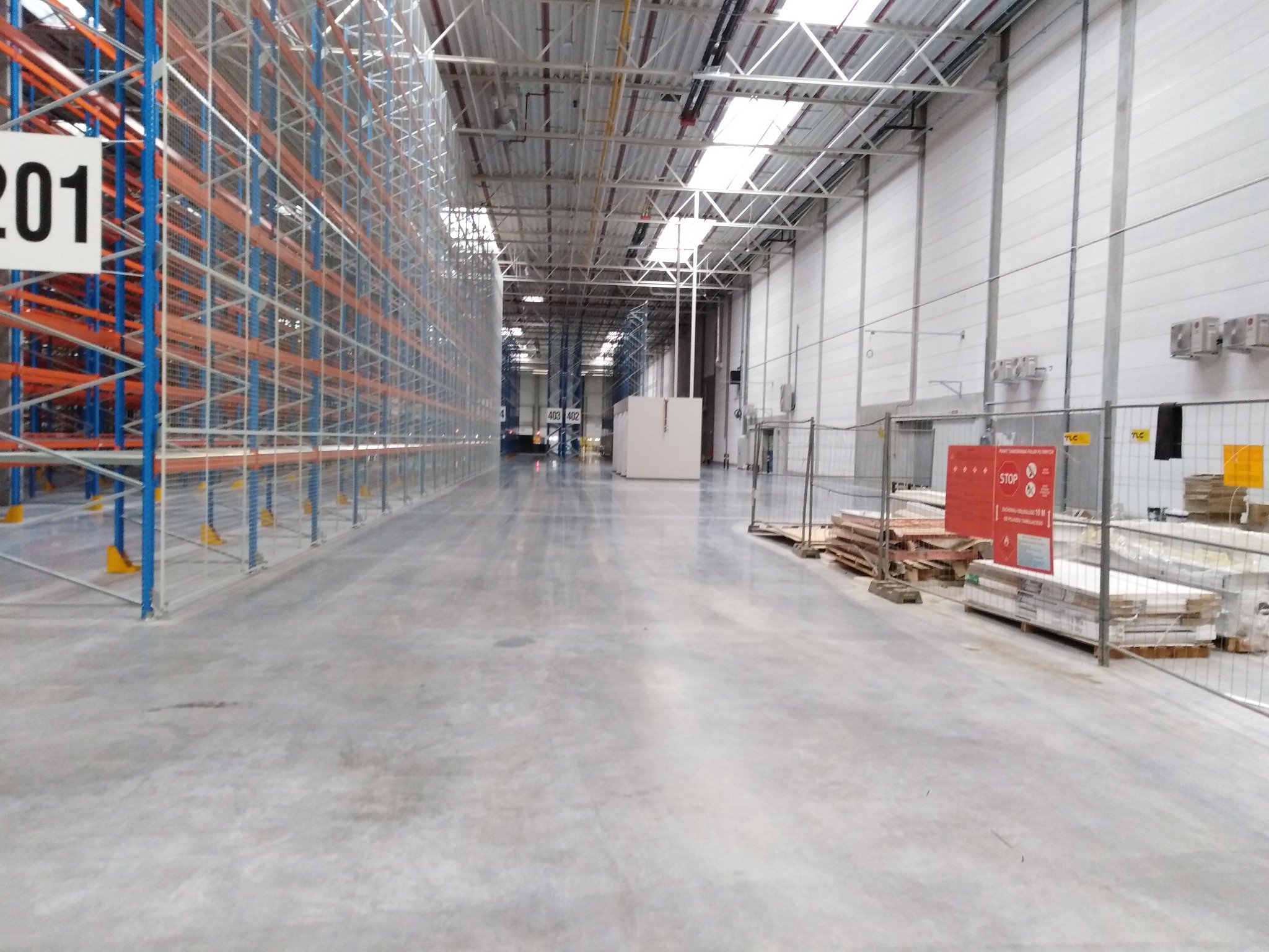 Pusta hala magazynowa z wysokimi regałami, numer 201 na białej tablicy, betonowa posadzka, widoczne materiały budowlane zabezpieczone siatką, sufit z widoczną konstrukcją, oświetlenie górne.
