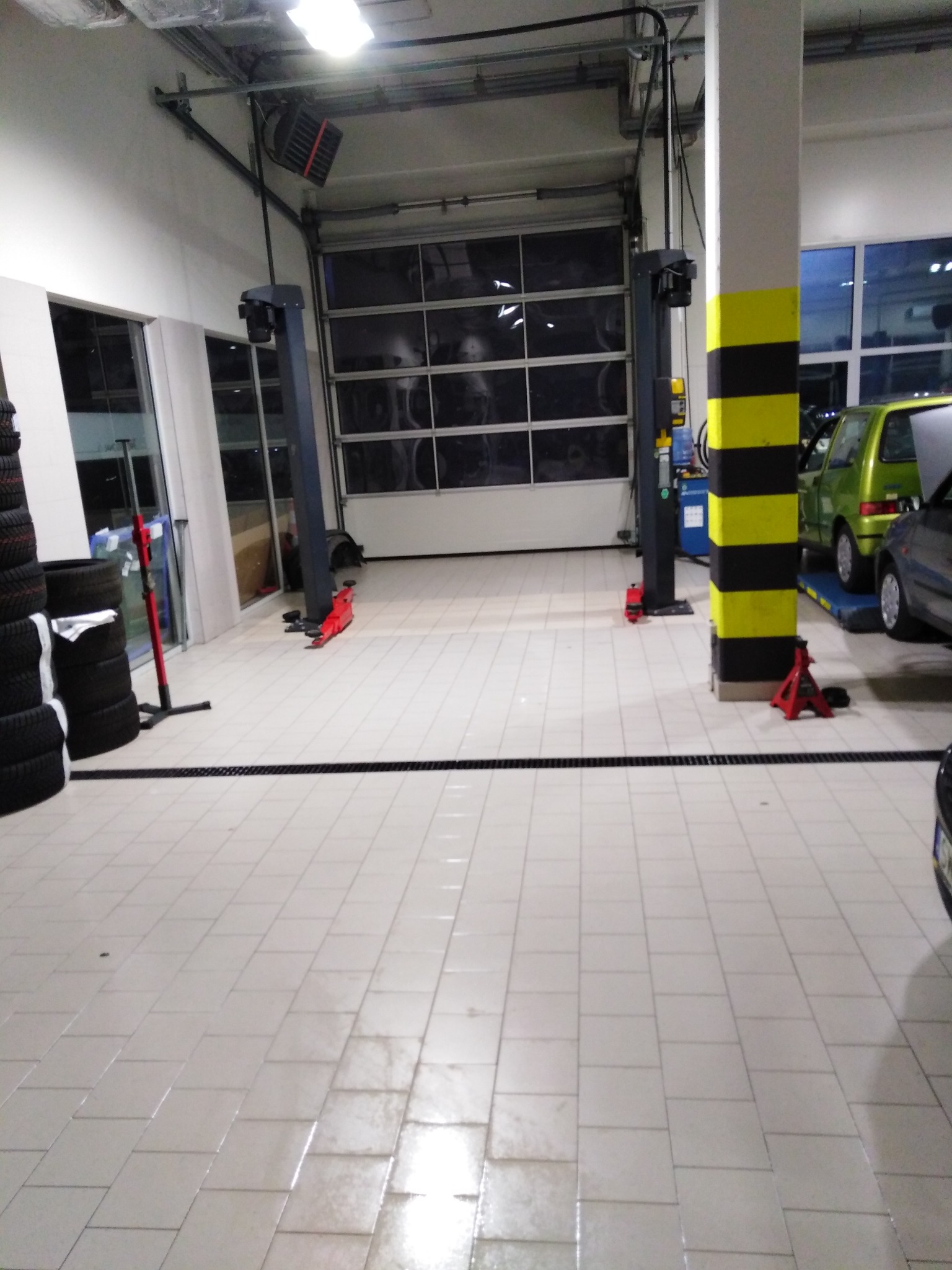 Czysta posadzka z płytek w warsztacie samochodowym z podnośnikiem dwukolumnowym i bramą segmentową w tle, obok stos opon i zielony samochód na podnośniku.